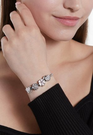 Silver bracelet featuring heart and flower charms with rhinestone accents. Worn on a wrist with a textured black top.