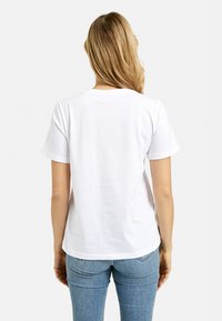 Blonde woman seen from the back wearing a plain white short-sleeve t-shirt and light blue jeans against a white background.