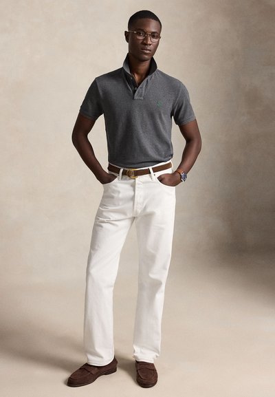 Homme portant un polo gris, pantalon blanc, ceinture marron, mocassins marron, montre et lunettes, debout, les mains dans les poches sur fond beige.