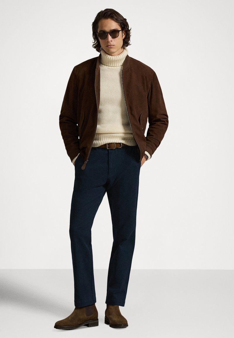 Giacca bomber in camoscio marrone sopra un maglione dolcevita color crema, abbinata a pantaloni blu navy e stivaletti Chelsea marroni. Occhiali da sole scuri completano il look.