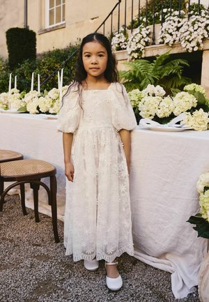 Ung pige i hvid blomstret blondekjole og hvide sko står på grus ved siden af et bord dekoreret med hvide blomster og stearinlys udendørs.