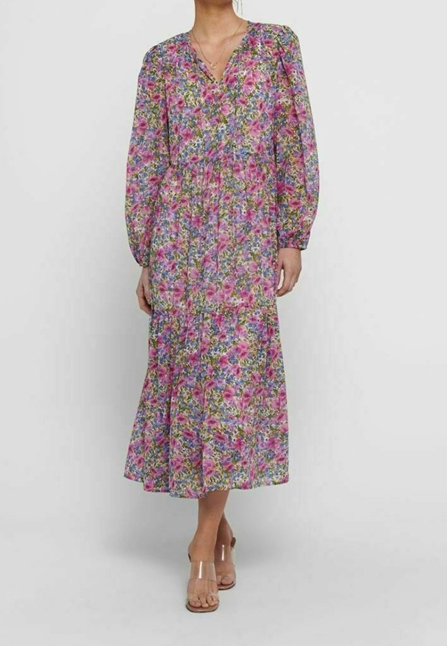 Femme portant une robe fleurie à manches longues, longue jusqu'aux chevilles, avec des motifs roses, violets et verts, associée à des sandales à talons avec brides transparentes.