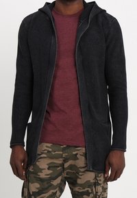 Cardigan noir texturé avec capuche, devant ouvert et deux poches avant, porté sur une chemise bordeaux et un pantalon camouflage.