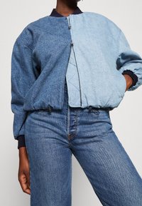 Jeansowa kurtka z podzielonym designem w ciemnoniebieskim i jasnoniebieskim odcieniu. Zawiera zamek błyskawiczny, prążkowany kołnierz oraz regulowany dół.
