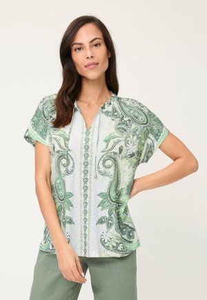 Donna con capelli castani lunghi che indossa una camicetta bianca a maniche corte con motivo paisley verde e pantaloni verde salvia, in piedi con la mano sui fianchi.