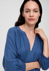 Chemisier bleu avec un col en V, des manches bouffantes froncées et des détails de couture visibles. Tissu doux avec une texture lisse.