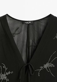 Schwarze Bluse mit V-Ausschnitt, versehen mit floralen Stickereien aus silbernen Perlen und einem zum Binden geschlossenen Taillenbund. Glattes Gewebe, leichtes Texture.