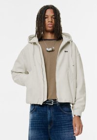 Lys beige zip-up hættetrøje med en løs pasform, parret med en brun og beige stribet skjorte. Mørkeblå jeans og et metallisk vedhængs halskæde.