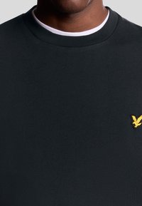 Zwarte katoenen sweatshirt met een ribbelnek en een klein geel arendlogo op de linkerkant van de borst. Soepele textuur, aansluitend ontwerp.