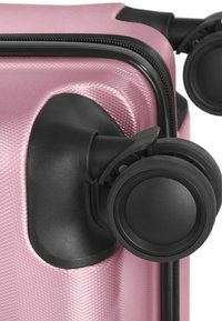 Coin d'une valise rigide rose texturée avec des roues doubles pivotantes noires et une fermeture éclair noire visible.