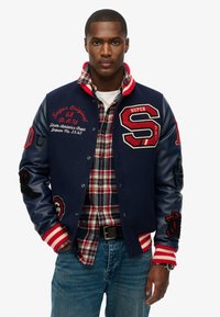 Chaqueta varsity azul marino con acentos rojos, mangas de cuero, logotipos bordados y puños a rayas. Llevada sobre una camisa a cuadros.