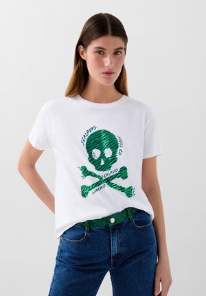 T-shirt bianca in cotone con un design di teschi e tibie a strisce zebrate verdi, accompagnato dal testo "SCALPERS", abbinata a jeans di jeans blu.