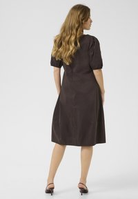 Rjava obleka do kolen s napihnjenimi rokavi, gladko teksturo in prilegajočim bodijem. Vključuje stranske žepe in enostaven dizajn.