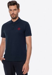 Námořnická modrá polokošile vyrobená z bavlny, se zapínáním na tři knoflíky, krátkými rukávy a červeným vyšitým logem na hrudi.