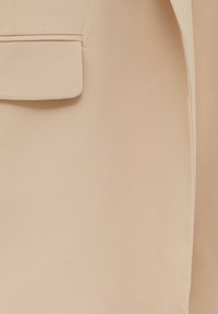 Détail de tissu de blazer beige avec une poche à rabat unique et des coutures visibles sur un matériau lisse et sur mesure.