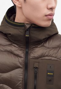 Gros plan d'une personne portant une veste matelassée marron avec des détails de fermeture éclair noirs et jaunes et un logo Blauer sur la poche poitrine.