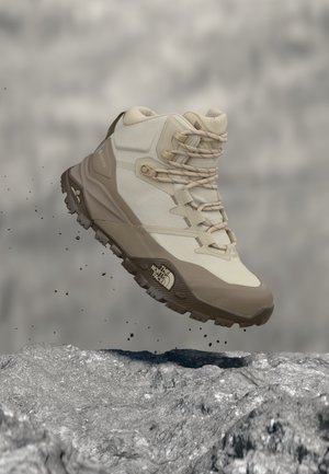 Béžová a hnedá turistická obuv so šnúrkami vo vzduchu nad sivou skalnatou zemou, ukazujúca robustnú podrážku a logo The North Face.