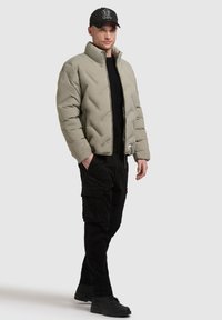 Uomo che indossa un giubbotto imbottito grigio chiaro, pantaloni cargo neri, un maglione nero, stivali neri e un cappellino nero con logo bianco, in piedi con le mani nelle tasche.