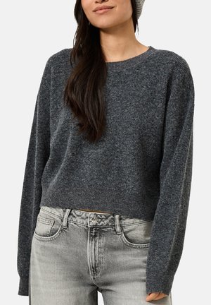 Vrouw draagt een donkergrijze gebreide trui, lichtgrijze gewassen spijkerbroek en een lichtgrijze beanie, getoond van borst tot heupen.