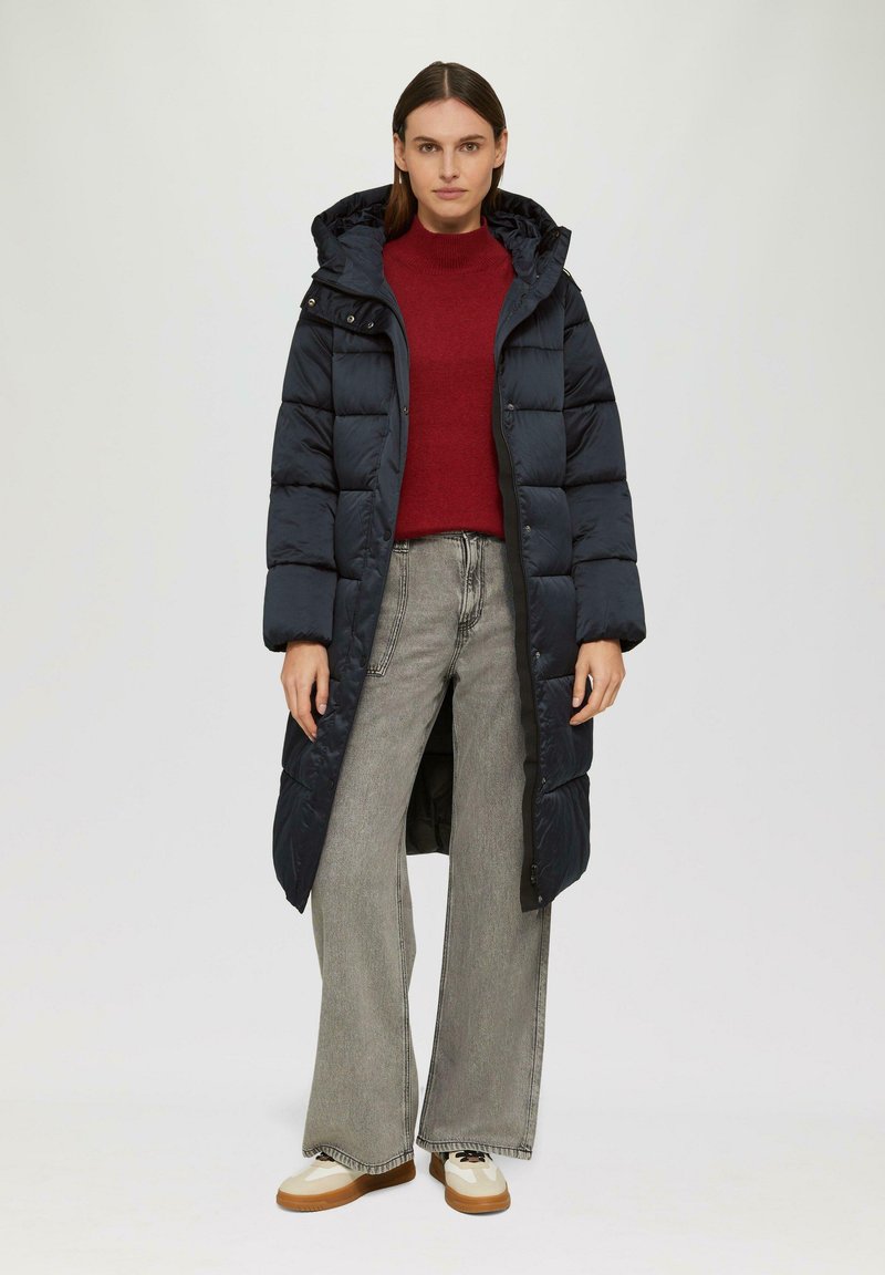 Marineblaue Steppjacke mit Kapuze, lang geschnitten und mit gestepptem Muster. Trägt man über einem roten Pullover und grauen, weiten Jeans.