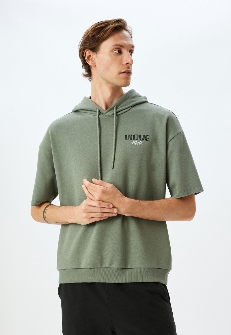 Zielona krótka bluza z kapturem z miękkiego materiału, z przednią kieszenią typu kangur, sznurkiem do regulacji kaptura oraz haftowanym logo "MOVE Master".