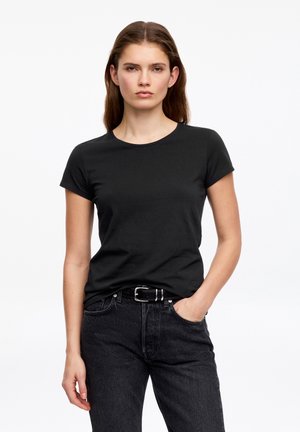 Kobieta o długich brązowych włosach, ubrana w zwykły czarny T-shirt włożony do wysokich czarnych dżinsów z czarnym paskiem, stojąca z jedną ręką w kieszeni.