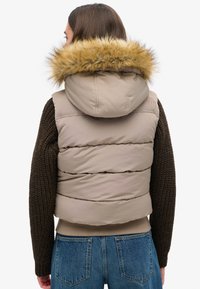 Beige gepolsterte Weste mit abnehmbarem Kapuze, ausgestattet mit braunen Strickärmeln und einem Kunstfellbesatz an der Kapuze für zusätzliche Textur und Wärme.