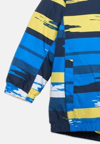 Värvikas tuulekindel jakk, millel on horisontaalsed triibud sinises, kollases ja valges. Valmistatud siledast kangast, millel on elastsed mansetid ja küljetasku.