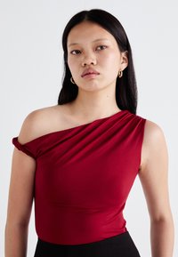 Top rosso scollato realizzato in materiale elastico, con design drappeggiato, texture liscia e silhouette aderente, abbinato a un fondo nero.