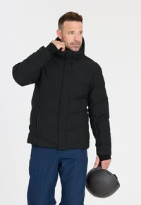 Schwarze isolierte Jacke mit Reißverschluss vorne, Kapuze und zwei Seitentaschen. Getragen mit blauen Hosen und hält einen grauen Helm.