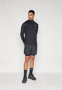 Uomo in outfit sportivo Nike nero in piedi con le mani dietro la schiena, che guarda a destra su uno sfondo chiaro e semplice.