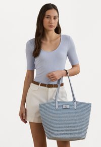 Gaiši zila austā somiņa ar divām rokturiem, ar strukturētu rakstu un taisnstūra logo uzlīmi priekšpusē.