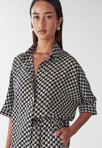 Košile s černobílým vzorem a geometrickými motivy, krátké rukávy a límec. Tkanina vypadá hladce a lehce.