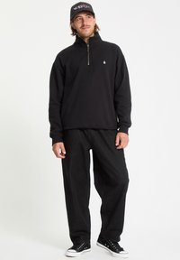 Pullover in pile nero con zip e un piccolo logo sul petto, abbinato a pantaloni neri larghi e sneakers nere.