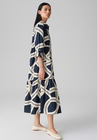 Long-sleeve, button-up dress in navy with cream abstract pattern, tiered skirt, soft fabric, and relaxed fit. Paired with light flats.
