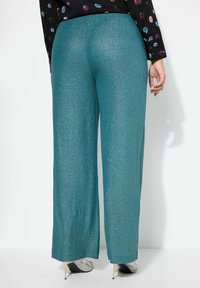 Pantalons évasés teal avec une texture scintillante, des poignets détaillés et des fentes latérales, associés à des talons aiguilles argentés sur fond blanc.