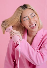 Brosse sèche-cheveux rose avec des poils, forme cylindrique, prise texturée et cordon électrique. Sur fond rose.