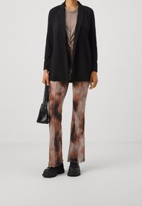 Blazer negro, top beige anudado, pantalones de campana en tonos marrón y crema con dobladillo ondulado, botas negras gruesas y un pequeño bolso negro.