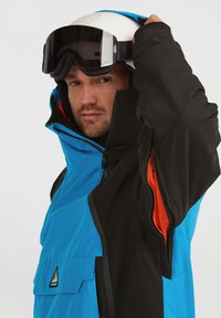 Chaqueta impermeable azul y negra con un alto cuello, bolsillos con cremallera y acentos de malla naranja en contraste, diseñada para deportes de invierno.