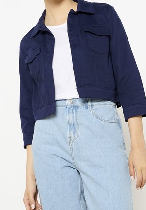 Przycięta granatowa kurtka z klapkowymi kieszeniami noszona na białym topie i jasnoniebieskich, wysokich jeansach na osobie stojącej.