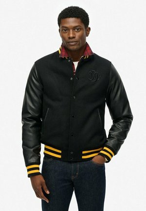 Chándal negro de estilo universitario con mangas de piel, rayas amarillas en el cuello y los puños, y un forro interior de cuadros. Cuenta con un emblema de marca en el pecho.