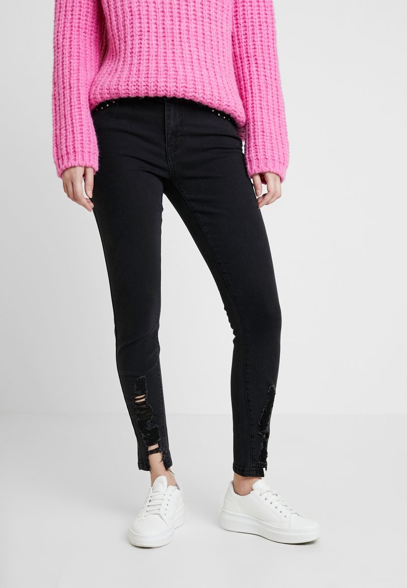 Persona vistiendo un suéter de punto rosa brillante, jeans ajustados negros rasgados y zapatillas blancas, de pie contra un fondo blanco liso.