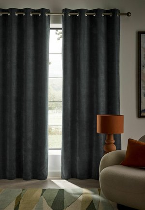 Dunkelgraue Vorhänge hängen teilweise offen an einem Fenster neben einem beigen, geschwungenen Sofa mit einem orangefarbenen Kissen und passender Tischlampe.