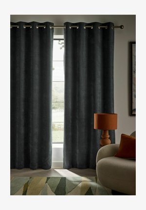 Dunkelgraue Vorhänge hängen teilweise offen an einem Fenster neben einem beigen, geschwungenen Sofa mit einem orangefarbenen Kissen und passender Tischlampe.
