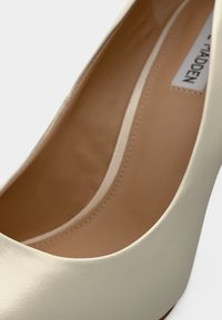 Chaussure à talon haut ivoire avec une surface lisse et texturée, bout arrondi et doublure intérieure beige. Coutures discrètes le long des bords.