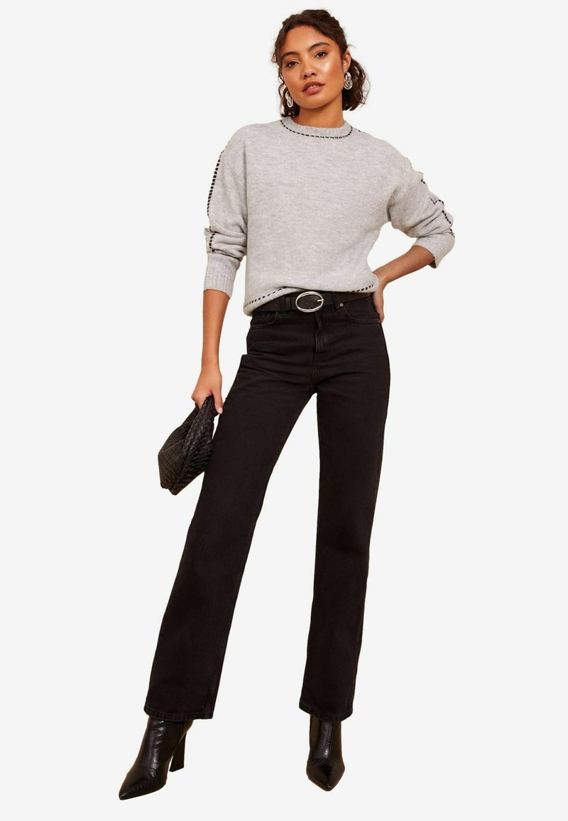 Grå sweater med sideskæring, parret med højtaljede sorte jeans og spidse hæle støvler, mens hun holder en vævet sort clutch.