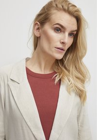 Helle beige Leinen-Blazer über einem rostfarbenen gerippten Oberteil. Der Blazer hat einen Reverskragen und eine lässige Passform. Haare in lockeren Wellen gestylt.