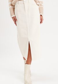 Falda midi de denim blanco con abertura frontal, cintura alta, bolsillos laterales y una textura sutil. Combinada con botines crema.