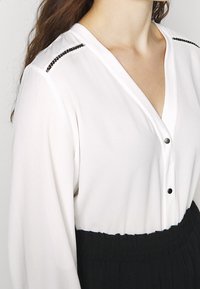 Un primo piano di una donna che indossa una camicia bianca con bottoni decorativi e una gonna nera, evidenziando i dettagli del colletto e delle maniche.