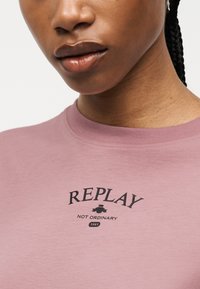 Κοντινή λήψη ενός ατόμου που φοράει ένα μωβ μπλουζάκι με μαύρο κείμενο "REPLAY", ένα μικρό γραφικό και τη φράση "NOT ORDINARY 1981" στο στήθος.